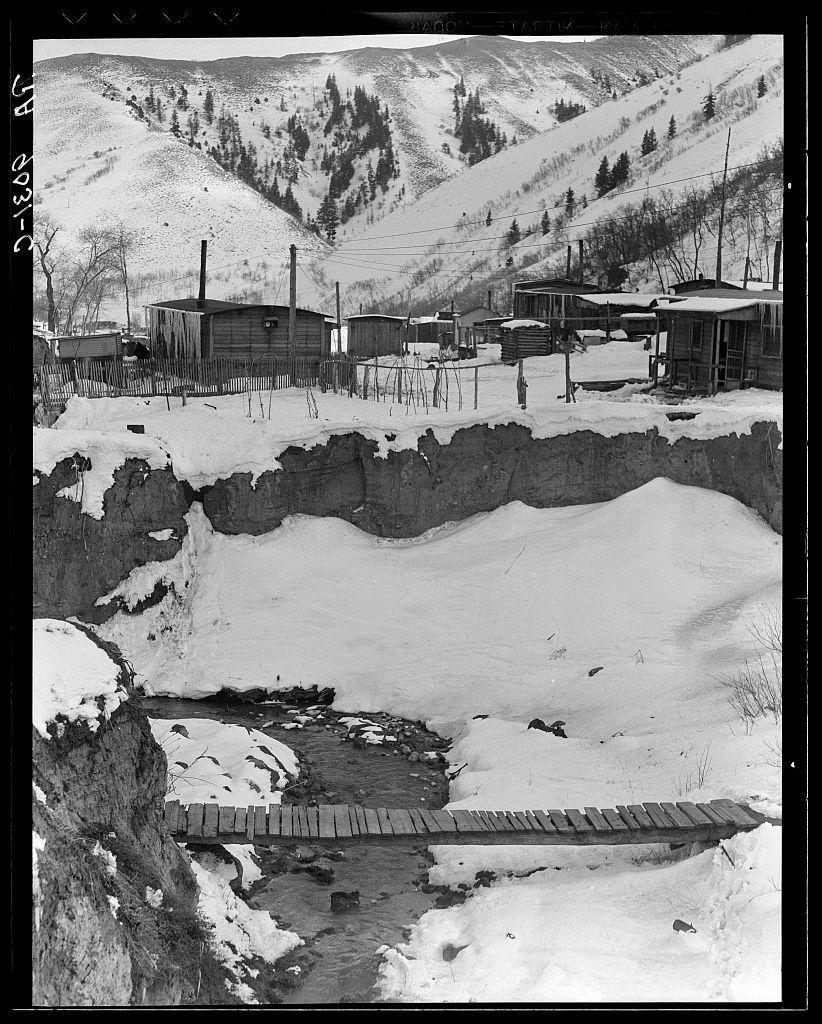 The image depicts a rural, wintery landscape with several wooden houses and outbuildings scattered across the terrain. Snow blankets much of the ground, indicating cold weather conditions. A small bridge constructed from logs spans over what appears to be a shallow stream or creek, partially covered in snow and ice. The backdrop features hilly slopes dotted with evergreen trees, suggesting this setting is within a mountainous region. Notable elements include wooden fences surrounding some properties, indicative of attempts at demarcation despite the spartan nature of the structures themselves.