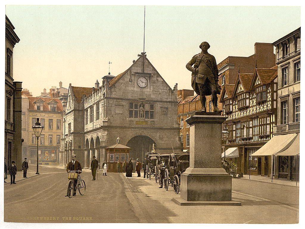 This image depicts a historical street scene in what appears to be the early 20th century. The focal point is an ornate clock tower with a figure of a man atop a pedestal, likely representing Sir Rowland Hill or another notable local dignitary from Shrewsbury's history. This structure serves as a grand entrance gateway marked "Shrewsbury," and beyond it lies what seems to be the town square bustling with activity.

Various individuals are seen walking on the pavement, some in period attire indicative of late Victorian-era fashion: men wearing suits or overcoats and hats, women dressed modestly. The streetscape is filled with historical architecture featuring Tudor-style gabled facades, timber framing, and ornate details typical for this era. There's a horse-drawn carriage on the right side while bicycles are also prominent, reflecting transitional times from traditional to modern transportation.

The scene suggests an everyday life in Shrewsbury during its heyday as "The Square," with shops lining one side of the street hinting at commerce and social interaction within this urban environment.