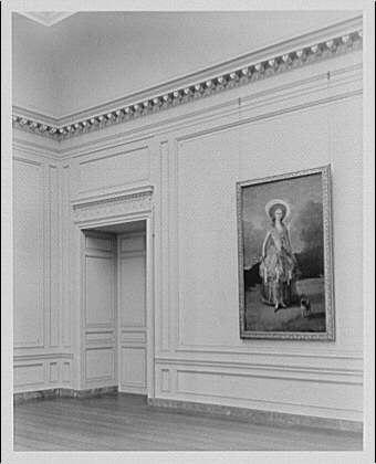 The image shows a black and white photograph of an empty room with classic architectural details, such as ornate cornices along the ceiling. On one wall hangs a framed portrait depicting a figure in historical attire standing on what appears to be a rug or carpet, holding a small object or piece of paper in their hand. The lighting suggests that this photo was taken before color photography became widespread and is likely from an early period given its monochromatic nature. There's no visible text within the image itself but references indicate it may have been part of an exhibit at the National Gallery of Art during a specific time frame, highlighting works by Horydczak, Theodor (1890-1971).