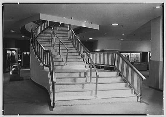 The image shows an interior view of a staircase with multiple levels, featuring white risers and steps. The stairwell has a modern design with metal railings that have alternating light and dark stripes for contrast against the concrete structure. Overhead lighting fixtures are visible on the ceiling, which appears to be tiled or paneled with recessed areas housing lights.

The surrounding environment suggests an indoor space with multiple levels connected by this staircase. The area seems spacious and well-lit, possibly part of a larger building such as a mall, office complex, or public facility. There is no visible activity on the stairs themselves; they are empty in the photograph.
<start of description>
This image depicts an interior setting featuring a prominent curved staircase with multiple levels