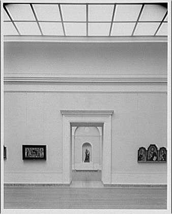 The image is a black and white photograph showing the interior of an exhibit room, likely from around 1920 to 1950. The architecture includes high ceilings with exposed beams or skylights providing natural light into the space.

In this section of the room are several framed art pieces displayed on both sides walls above pedestals. On the left side wall is a framed painting titled "Pezzana," and next to it, there's another artwork without visible text due to angle and resolution constraints in the photograph. The right-side wall displays two other artworks or artifacts with no discernible titles.

In front of the central opening on one end, which serves as an entranceway into a different room, is a statue placed upon what appears to be a pedestal. This display area stands out from the rest due to its positioning and lighting accentuation.

The overall composition suggests that this exhibit was part of a larger gallery or museum with specific arrangements for displaying artworks within historical context or thematic coherence.