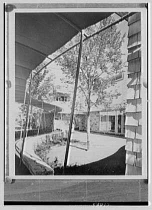 The image is a black and white photograph of an outdoor courtyard area, featuring trees with leafy branches. The photo appears to be taken from the inside looking out through what seems like a screened or netted fence that partially obscures the view. There are buildings visible on both sides of the courtyard. One building has large windows and siding made up of rectangular panels. A walkway curves around the center of the area, with benches placed alongside it for seating. The overall atmosphere suggests an open-air shopping arcade or a similar recreational space within urban architecture from 1948 in Scarsdale, New York.