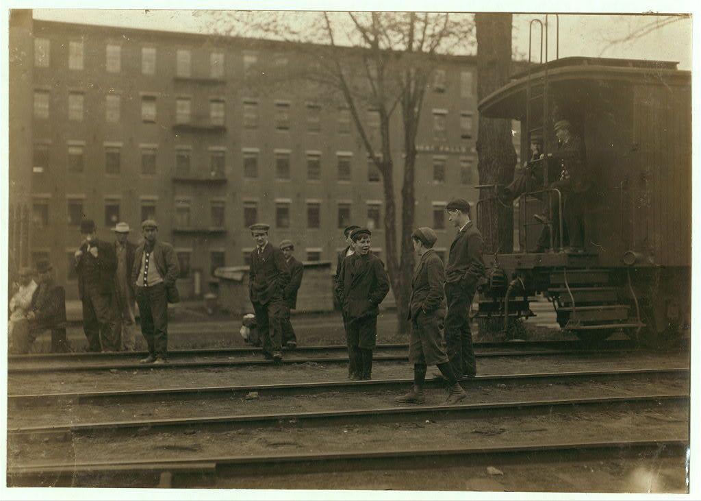 The image is a black and white historical photograph depicting several individuals standing on railway tracks, likely during the early 1900s. The scene appears to be an industrial or working environment given the presence of trains in the background and what seems to be workers dressed in various attire indicative of different roles or ranks within this setting.

Several men are wearing suits with ties, hats, and one is holding a newspaper, suggesting they might be supervisory staff or possibly off-duty individuals taking a break. Others appear more casually dressed, indicating they could be laborers at the Great Falls Manufacturing Company mentioned in the caption. There's an older man standing on top of what appears to be railway carriages looking down towards those below.

In the background, there is a large multi-story industrial building with numerous windows and doors, typical of factory or mill architecture during that era. The environment looks cold as suggested by the attire of individuals wearing heavy coats and hats suitable for winter weather conditions in New Hampshire on May 19th as indicated by the caption. There's also sparse vegetation visible which reinforces a colder climate.

The photograph is credited to Lewis Hine, known for his work documenting child labor during that period through the National Child Labor Committee, though this specific image does [...]