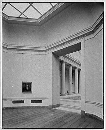 The image depicts an interior view of a spacious and elegant gallery room within the National Gallery in Washington, D.C., during the early to mid-20th century. The photograph is black-and-white, highlighting the architectural features such as classical columns lining one side and a high ceiling with skylights that allow natural light into the space.

On the wall opposite the entrance stands an oil portrait encased within a simple frame. Below it lies two long, narrow vents or air ducts on the wooden floor, which appears to be polished wood given its reflective sheen.

The overall atmosphere exudes a sense of grandeur and historical significance, evoking the ambiance typically associated with classical art museums during that era. The absence of people in the scene underscores the quiet contemplation often encouraged within such settings for visitors appreciating fine arts.