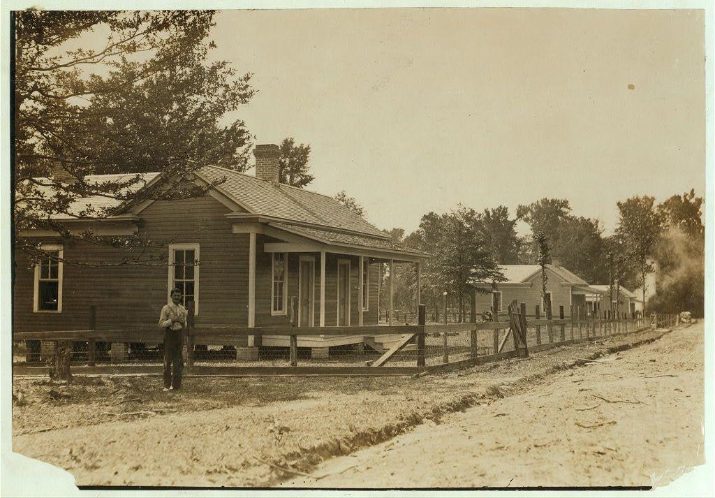 The image is a sepia-toned photograph depicting a rural scene from the early 1900s. A two-story wooden house with a wrap-around porch and multiple windows stands prominently in the foreground, surrounded by what appears to be sparse vegetation or grassy area. The architecture suggests an older American home design, possibly indicative of late 18th-century construction methods.

A fence encloses part of the property where several individuals are visible: one person standing at a distance on the left side of the image and another seated near the middle-right section within what looks like a wooden structure or shed. The sky is overcast with no discernible clouds, hinting that it might be an early morning or late afternoon setting.

The landscape extends into the background where we can observe other similar houses in close proximity to each other on both sides of a dirt road, suggesting this may be part of a small community housing area for workers. The presence of smoke rising from one building indicates ongoing activities within these homes.

Overall, it portrays an idyllic yet modest living environment with no signs of modern amenities or luxury elements. It captures the essence of simple rural life possibly tied to labor in nearby mills as mentioned in additional information about this image indicating working conditions being good for a mill settlement at Magnolia, Mis [...]