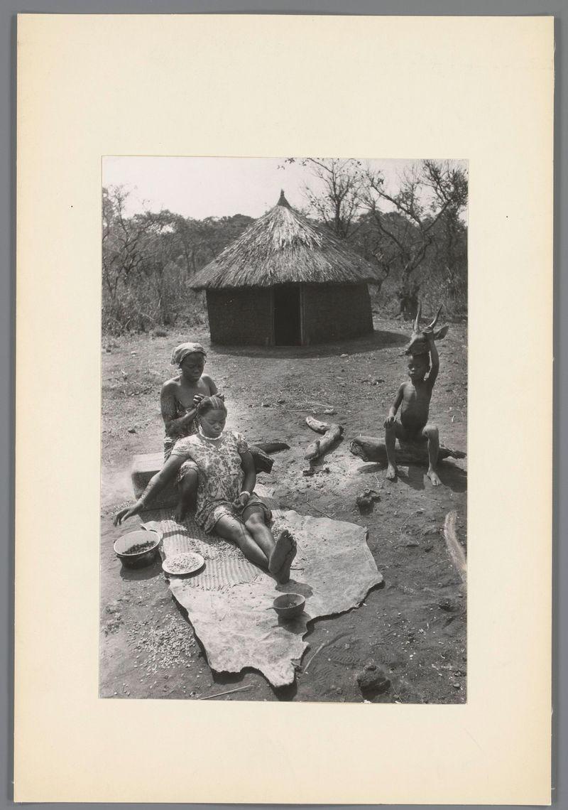 The image depicts a rural scene with three individuals engaged in various activities. In the foreground, there is a woman seated on what appears to be animal skin spread out on the ground, wearing patterned clothing and possibly engaging in food preparation or sorting of items from a bowl next to her. Beside her lies another large flat object that could serve as a place for drying goods.

In the center background stands an individual with their hands raised, holding horns above their head which suggests they are interacting with an animal's skull, likely performing some sort of ritualistic act or examination.

The third person is partially obscured and appears to be walking away from the camera toward the thatched-roof hut in the upper left corner. The surroundings include sparse vegetation, indicative of a dry climate, and there seems to be no other significant structures nearby beyond the small wooden hut with its straw roof which serves as a focal point for the scene.

This photograph captures moments that reflect daily life or traditional practices within this community setting, providing insights into their customs and environment.