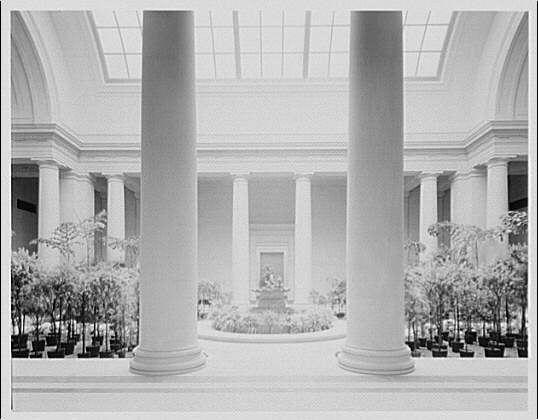 This black and white photograph captures the grandeur of a spacious indoor garden court, likely part of an art museum or gallery. The space is dominated by two large classical columns that frame the scene symmetrically on either side. These columns are set against a backdrop of tall windows with glass panes allowing natural light to flood into the area.

In front of these columns lies a meticulously maintained lawn surrounded by rows of potted plants, creating a neat and orderly pattern across the expanse. The central feature is an ornate fountain or sculpture that serves as a focal point in the court's design. Beyond this centerpiece, more vegetation can be seen extending into what appears to be another section of the garden.

The overall atmosphere conveyed by the photograph is one of serenity and elegance, with careful attention paid to both architecture and horticulture within the space. The absence of color emphasizes textures and contrasts, drawing focus to details such as the smooth surfaces of the columns, the varied foliage of plants, and the reflections on any water in the fountain.

The image provides a glimpse into an environment designed for contemplation and appreciation of art and nature, reflecting the design sensibilities prevalent during its time period. The photo's provenance is linked with "National Gallery" which suggests that this space may be part o [...]