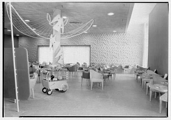 The image depicts an interior scene of a restaurant named the Bird Cage, dating from February 5, 1949. The establishment appears to have a modern and somewhat eclectic design with large tables arranged for dining. A notable feature is a whimsical chandelier resembling antlers or branches, suspended above the center of the room.
The walls are adorned with what seems like textured wallpaper or paneling in varying shades, creating an interesting backdrop that adds visual interest without overwhelming the simplicity of the space. The flooring appears to be a light-colored material, possibly linoleum or vinyl, which was common for such establishments at the time due to its durability and ease of cleaning.
The photograph is rendered in black and white, suggesting it may have been taken with an older camera that used film rather than digital technology. There are no people visible within the frame; this could indicate a moment when the restaurant was not open or possibly captured for promotional purposes prior to opening hours. The lighting seems even throughout the room, likely from overhead fixtures out of view in the photograph.
The image is identified as part of a collection by Gottschol-Schleisner Inc., potentially indicating their role either as photographers or an organization involved with documenting this particular business establishment during that era.