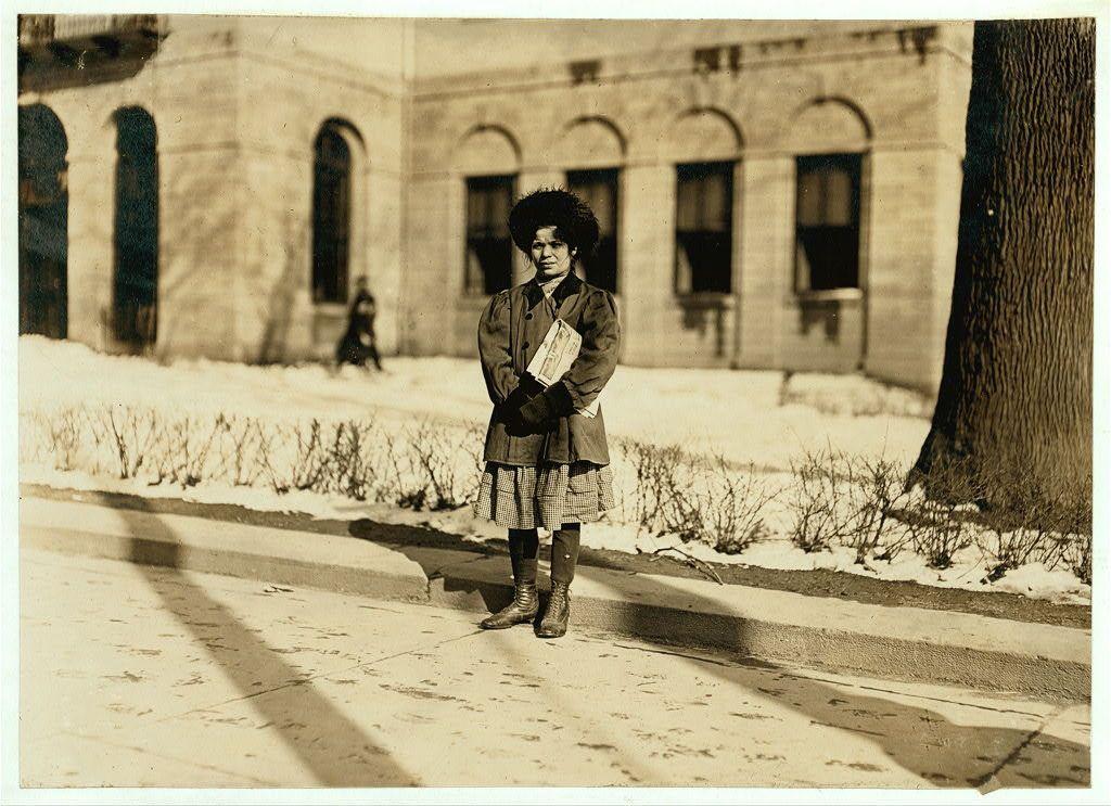 The image is a sepia-toned photograph depicting an individual standing on what appears to be a sidewalk, with buildings and leafless shrubbery in the background. The person is wearing dark boots, patterned tights or leggings, knee-length shorts or skirt-like pants, and gloves, suggesting cold weather conditions. They are holding something under their arm that could possibly be papers or tickets, hinting at some form of work being performed by this individual.

The setting seems to be an urban environment with architectural features typical of early 20th-century American cities. The person's clothing style is modest and practical, suitable for a child engaged in laborious activity during the time period indicated by their attire. There are no explicit indicators of gender or race that inform our interpretation; however, it should be noted that historical images like this were often taken from an era where such distinctions may have been less overtly considered.

Additional context provided suggests that Mary Dunn was 11 years old at the time and had been selling for one year. The mention of Hartford, Connecticut indicates a specific location which could aid in dating the photograph further.