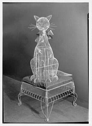 The image depicts a wire sculpture of a seated cat, which appears to be made from metal wires. The structure is placed on top of what seems to be an ornate bird cage or similar object with intricate legs and detailing.
The photograph itself is in black and white format, indicating it may have been taken during the mid-20th century, as suggested by the caption mentioning 1949 Feb. It's important to note that this image was found through a search for Lord & Taylor business records held at Gottschalk-Schleisner Inc., which is associated with an archive of negatives and photographs dating from around January in New Jersey.
For those who are visually impaired, the sculpture captures attention due to its unique design. The cat's posture appears relaxed as it sits comfortably on top of a seemingly delicate object made for birds' safety or containment purposes.