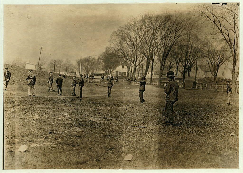 The image depicts a sepia-toned photograph of several people playing baseball on an open field. There are around ten individuals, mostly male and dressed in early 20th-century clothing such as hats, coats, and trousers. The players appear to be engaged in various stages of the game: one is throwing a ball, another appears to have just caught it, while others stand or walk casually on the grassy field.

In the background, leafless trees line what seems like an urban area with buildings visible across the horizon under a cloudy sky. A prominent hillside can be seen in the far distance and there are some wooden structures, possibly fences, indicating property boundaries within this setting.

The photograph is labeled "Noon hour ball-game at Quidwick Co. Hill, Anthony, R.I." It notes that all individuals work in the mill mentioned; however, specific details about their occupations or roles cannot be discerned from the image alone. The location specified is Anthony, Rhode Island.

Given its historical context and subject matter, this photograph could offer valuable insights into leisure activities during a particular era as well as provide information on working-class recreation spaces within urban environments of that time period in New England.