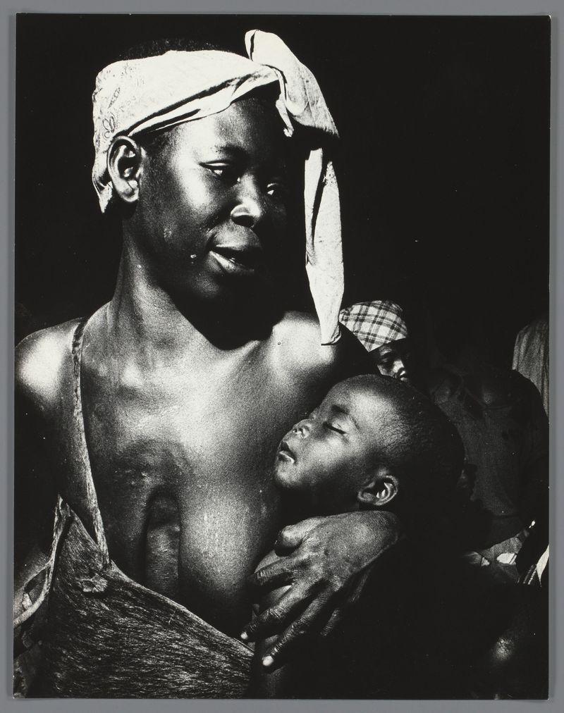 The image is a black and white photograph depicting two individuals, an adult wearing what appears to be traditional African attire with draped clothing wrapped around their body. This person has a towel or cloth head wrap tied loosely at the back of their neck, revealing one ear prominently in profile view. The individual's gaze is directed toward something out of frame on the left side, and there are distinct marks visible across the chest area.

In close embrace to this adult figure is a younger child with round cheeks and open eyes looking slightly upwards or off into space. The child appears contemplative and seems to be resting against the older person's torso for support or comfort.

The background of the photograph fades into darkness, focusing all attention on these two figures in stark contrast. No discernible context or setting is provided within this image; it captures a moment that feels intimate yet open-ended regarding its circumstances or story.

This particular photo does not provide any additional information beyond what can be inferred from the visual content itself, and without further knowledge of Ed van der Elsken's "Vrouw met kind tijdens een dans" context.
