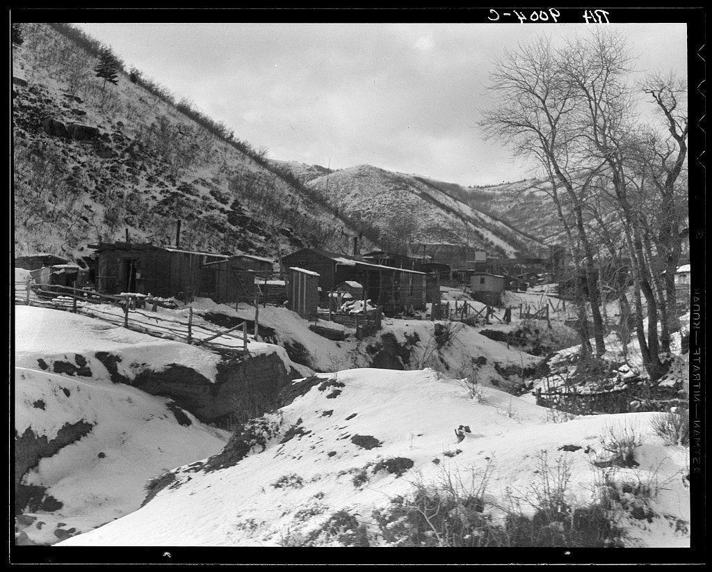The black and white photograph depicts a stark rural landscape, likely from an early to mid-20th-century setting. The scene is characterized by snow-laden slopes that descend into a cluster of simple wooden structures with gabled roofs, indicative of modest dwellings or outbuildings nestled on the hillside. Bare trees dot the barren terrain around these buildings, suggesting it's winter due to the undisturbed snowy landscape and leafless branches. The photograph captures an industrial aspect too; partially buried in snow is a large cylindrical structure resembling mining equipment. This setting suggests this image could be related to coal mining activities near Price, Utah as referenced by "Consumers" which may allude to Consumers Energy Company that operated mines in the region during its history of mining operations between 1905 and approximately when it closed down after World War II due to economic decline.