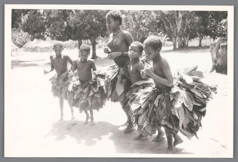 The image depicts a group of children engaged in what appears to be a cultural or celebratory dance. Four young boys are wearing traditional skirts made from large, flat leaves, possibly banana leaves, wrapped around their waists and secured with ties at the back. They have bare chests and legs, suggesting warm weather conditions typical for tropical environments.

The boy on the far left is in motion, perhaps dancing, as indicated by his raised arms and bent knee. The second child from the right looks towards him, possibly following along or waiting to move next. All four boys are holding what appears to be a cup or container of some sort, which they might use during their dance.

Behind them stands an adult woman with her hands on hips, observing the children's activity. She has short hair and is dressed in casual clothing that doesn't stand out but suggests practicality for outdoor activities.

The background features trees providing shade and what could be huts or structures indicative of a rural settlement. The setting looks like it might be part of an open-air community space with ample sunlight illuminating the scene, casting sharp shadows on the ground.

This photograph evokes a sense of cultural expression among young individuals in their traditional attire, captured during daytime activities that may hold significant meaning within their culture or local customs.