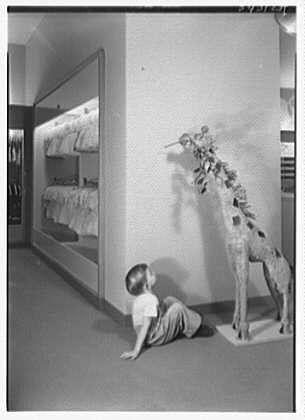 The image depicts a young boy on his knees looking up at an intricate wire sculpture of a giraffe with multiple figures attached to it. The scene appears to be inside a store, possibly Lord & Taylor in Millburn, New Jersey as indicated by the caption provided. There is also what looks like a display case or shelving unit containing various items above and beside the boy which adds depth to this composition. The image has a vintage quality with its black-and-white tone and appears to be taken on February 5th,1949 according to context given in Lord & Taylor business records.
