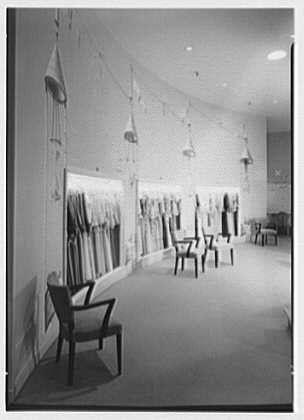 A vintage black and white photo of a clothing store, likely from the 1940s. The storefront features mannequins displaying various outfits in rows along shelves on one side and hanging garments above them. A modern chair is placed near the window display, adding contrast to the otherwise traditional retail space.