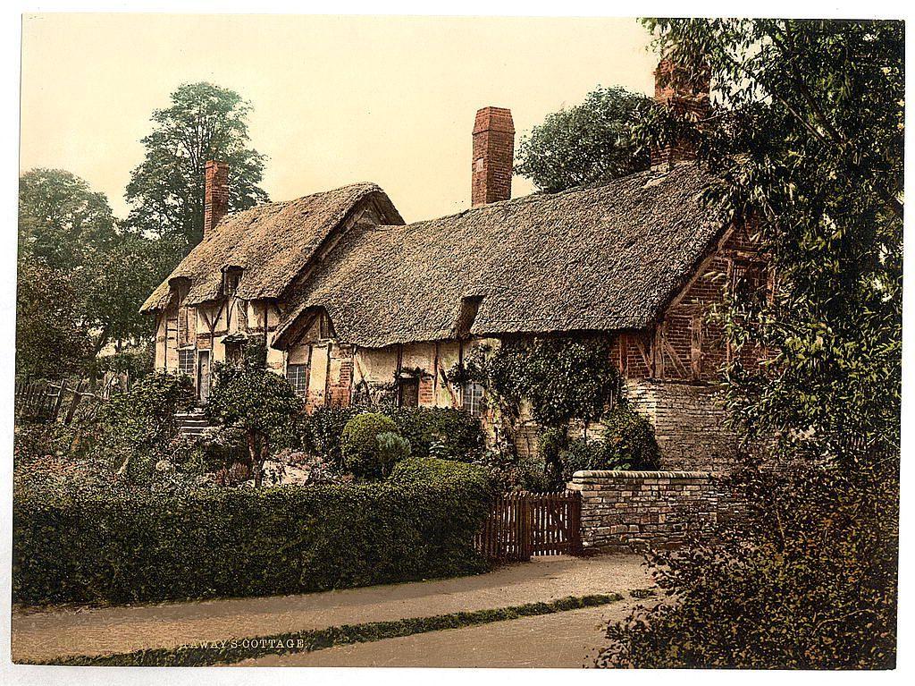 This image shows a traditional thatched cottage with exposed timber framing, indicative of Tudor architecture. The house features two prominent brick chimneys and is surrounded by lush greenery including hedges, bushes, and trees. A wooden fence borders the property, leading to what appears to be an entrance gate partially obscured by foliage. Stone walls are visible in various parts around the cottage's perimeter, suggesting a rural or semi-rural setting. The photograph has a vintage look with sepia tones, hinting at its historical significance as it is labeled "Ann Hathaway's Cottage," possibly referencing William Shakespeare’s birthplace located on Henley Street in Stratford-upon-Avon, England. This image captures the essence of historic English countryside and is part of a collection from around 1890 to 1900 by loener.nl