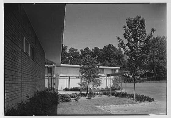 This black and white photograph depicts an entrance view of a building from August 23, 1949. The image features two distinct structures: the first is a dark brick facade with large windows on its upper level, while the second appears to be a lighter-colored structure possibly made of metal or concrete, situated adjacent to it and featuring a flat roof.

The foreground shows a well-maintained garden area with neatly trimmed bushes, shrubs, and small trees. There's also an open space that seems like a parking lot or pedestrian walkway leading up to the buildings. The overall scene is framed by tall greenery in the background indicating a suburban setting surrounded by trees.

Given additional information about Lord & Taylor business in Millburn, New Jersey, it suggests this building could be associated with commercial activities from that period and location. Gottscho-Schleisner Inc., mentioned as well, might refer to an architectural or photography company related to the creation of the image; however, without further context, their exact role remains uncertain.

The overall composition emphasizes modernist design principles common in mid-20th century architecture with its clean lines and functional aesthetic.