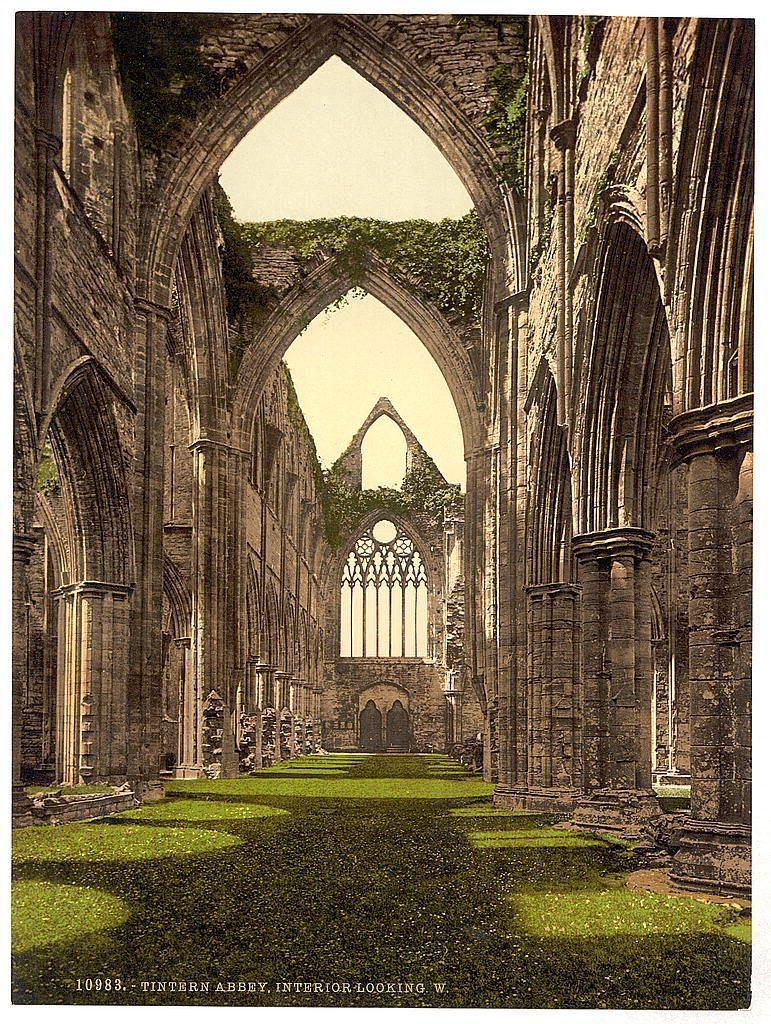 The image portrays an opulent and grandiose abbey interior, specifically Tintern Abbey in England. It features a long nave or central part of the building with intricately designed stone arches that reach towards a vaulted ceiling at both ends. The walls on either side are also adorned with large windows filled with intricate tracery, allowing ample light to flood into the space and illuminate green mossy floors below. Overgrown ivy clings onto parts of the ruins, adding an element of nature reclaiming human-made structures over time.

The Abbey's architecture is reminiscent of medieval Gothic style known for its height, pointed arches, ribbed vaults, and flying buttresses, although those are absent in this ruinous depiction. The sunlight casts warm hues on the stone surfaces, highlighting textures like moss growths and weathered stonework details. The composition exudes a sense of historical reverence and suggests that Tintern Abbey was once an imposing structure with significant spiritual or communal importance.

There's no visible activity within the abbey; it is deserted except for what appears to be ruins left behind by time, suggesting the building has seen better days but still retains much of its former grandeur. The absence of people may also emphasize solitude and reflection in this historic site.