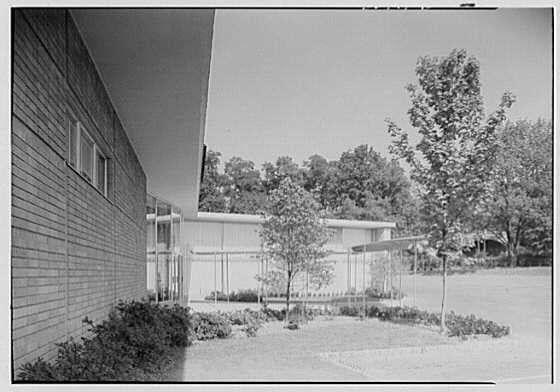 The image shows a black and white photograph of an outdoor setting, featuring two buildings with modern architectural designs. The building on the left appears to be made of wood paneling while the one on the right is constructed with what looks like concrete or stone materials. In front of these structures are well-maintained gardens containing shrubs, bushes, and trees that add a touch of greenery.

The photograph captures an entrance view from an angle where we can see both buildings prominently framed by natural elements. The sky in the background suggests it is daytime with no visible clouds or weather disturbances.

This image appears to be related to Lord & Taylor business located in Millburn, New Jersey as mentioned in a caption or description associated with this photo. It was taken on August 23rd, presumably during the year when Gottscho-Schleisner Inc., possibly an architectural firm known for producing such photographs of buildings and structures.

The URL provided indicates that further information can be found at https://images.loener.nl/gottscho-schleisner/full/691e/691efe89450e57a267500a10.jpg, which might lead to more details about this photograph and its historical context.