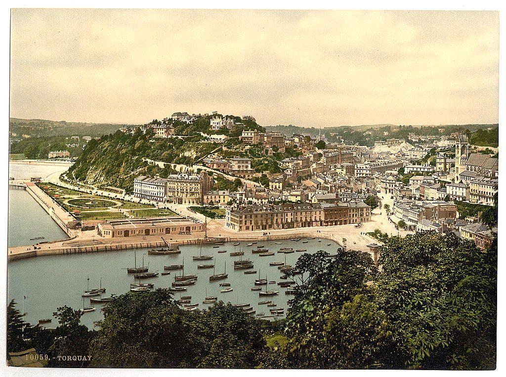 The image depicts a bustling seaside town situated on elevated terrain overlooking the harbor. The scene is dominated by a mix of residential and possibly institutional buildings, with their roofs showing various designs such as dormer windows and steeply pitched sloped rooftops indicative of older architectural styles. A prominent church or chapel can be identified towards the center-right part of the image due to its tower structure rising above the surrounding edifices.

The town is built on a hillside that descends toward a dock area filled with numerous boats, including what appear to be rowboats and small sailing vessels indicative of leisure activities. The harbor itself extends into the distance where it meets open water or possibly another body of land extending beyond the image's view.

In the foreground, lush greenery is visible, suggesting well-maintained parks or gardens along the waterfront which adds a touch of natural beauty to the urban setting. Above all this activity lies an expansive sky with scattered clouds, providing a serene backdrop for the busy scene below.

The image carries historical significance as it appears to be a vintage photograph from Torquay in England during approximately 1890-1900, capturing life and architecture of that era within Britain's South West region.