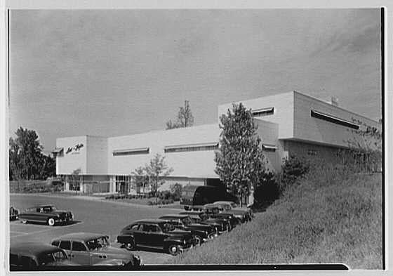 The image displays a black and white photograph of the exterior view to the south facade of Lord & Taylor's business in Millburn, New Jersey. This was taken on August 23rd, 1949 by Gottscho-Schleisner Inc., which is noted as the photographer for this work.
In the image, we see a large building with modernist architectural features characteristic of mid-20th-century commercial design. The structure has a flat roof and clean lines, suggesting it's likely from that era. It sits in an expansive parking lot filled with vintage automobiles indicative of the time period being depicted. Several cars are parked directly outside the entrance to the business.
The surrounding environment includes some greenery, like trees and grassy areas, which add context for the building's location within a suburban setting. The monochrome nature of the photograph adds historical atmosphere, while also emphasizing architectural details over color variations.
There is no visible signage or branding that would indicate what specific services Lord & Taylor offered at this time; however, it can be inferred as part of its business identity during this period based on its prominence and architecture style.