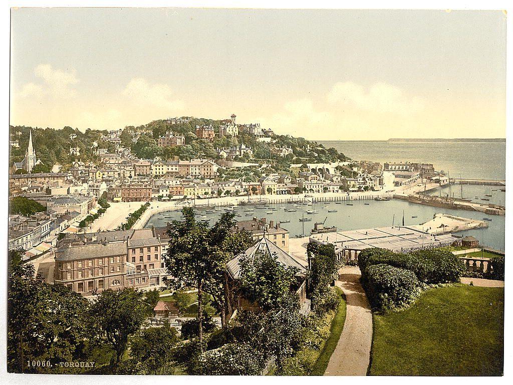 The image depicts a picturesque coastal town, likely captured during the late 19th to early 20th century given its vintage style. The scene is viewed from an elevated vantage point overlooking Waldron Hill in Torquay, England. The foreground features lush greenery and a well-maintained garden pathway leading towards a row of bushes on both sides, suggesting careful landscaping.

In the middle ground, there's a prominent waterfront with several buildings housing docks where boats are moored. Several ships or vessels can be seen anchored within this marina-like area. A mix of residential and commercial architecture lines the harbor-side streets, including large houses with multiple stories and rows of smaller structures that could serve as shops or apartments.

The background reveals Torquay town spreading across a hillside with an assortment of buildings varying in size, height, and architectural style. The dense arrangement indicates significant urban development. Among these constructions stands out several larger edifices, possibly churches, given their towering spires and classical features such as steeples and domed roofs.

The image's color palette is rich yet muted, indicating the use of early photographic techniques like albumen silver prints or similar processes common at that time before modern chemical processing. The sky above appears partly cloudy but allows e [...]