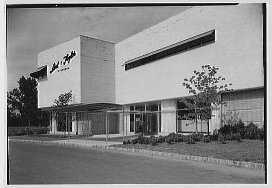 The image is a black and white photograph depicting the entrance facade of Lord & Taylor's business in Millburn, New Jersey. The structure features clean lines with a flat roofline on one side while an elevated area extends from it forming part of what appears to be two connected buildings that make up this retail establishment. It has large glass windows which seem well-ventilated and accessible for pedestrians by wide doors or entrances leading into the complex; no human figures are visible in view, suggesting a quiet moment outside at dusk with clear skies above as indicated by absence of shadows on building surface itself.

There's greenery surrounding it - grassy areas along the sides, neatly trimmed bushes bordering pathways to front entrance and perhaps young trees planted for aesthetics. The parking lot is paved but empty from what can be seen in this shot that captures only part of a street alongside with no moving cars or people visible indicating either quiet hours at dusk or perhaps an off-peak time not busy during working day.

The company's name "Lord & Taylor" is prominently displayed on the building, reinforcing its identity and providing context for who owns this establishment. The photograph was taken by Gottscho-Schleisner Inc., a noted photography firm in New York City known to have captured many important urban landmarks among other subjects during va [...]