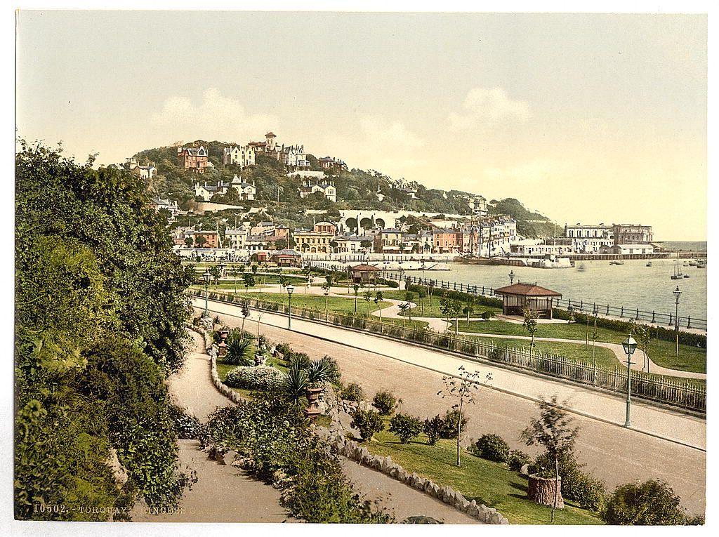 The image displays a picturesque and historical view of Torquay, situated along the coast in England. This scenic panorama captures an elevated vantage point overlooking a bustling town nestled on a hillside with various buildings indicative of early 20th-century architecture. These structures range from residential homes to larger edifices possibly serving commercial or administrative purposes.

Adjacent to the urban landscape is a well-maintained promenade, characterized by neatly paved paths, vibrant greenery including shrubs and trees, ornamental bushes along curving hedges, lampposts, benches for leisurely strolls, and flowerbeds adding splashes of color. The promenade appears tranquil yet inviting, likely serving as a popular spot for both locals and tourists.

Beyond the immediate foreground lies an expansive body of water that extends towards the horizon where it meets distant buildings suggestive of industrial or maritime use. This waterfront setting implies Torquay's significance in trade or shipping during its heyday.

The overall aesthetic is reminiscent of late Victorian to early Edwardian eras, suggested by architectural styles and urban planning typical of those periods. The image evokes a sense of calm elegance, characteristic of the British seaside towns that flourished as tourist destinations during this era.