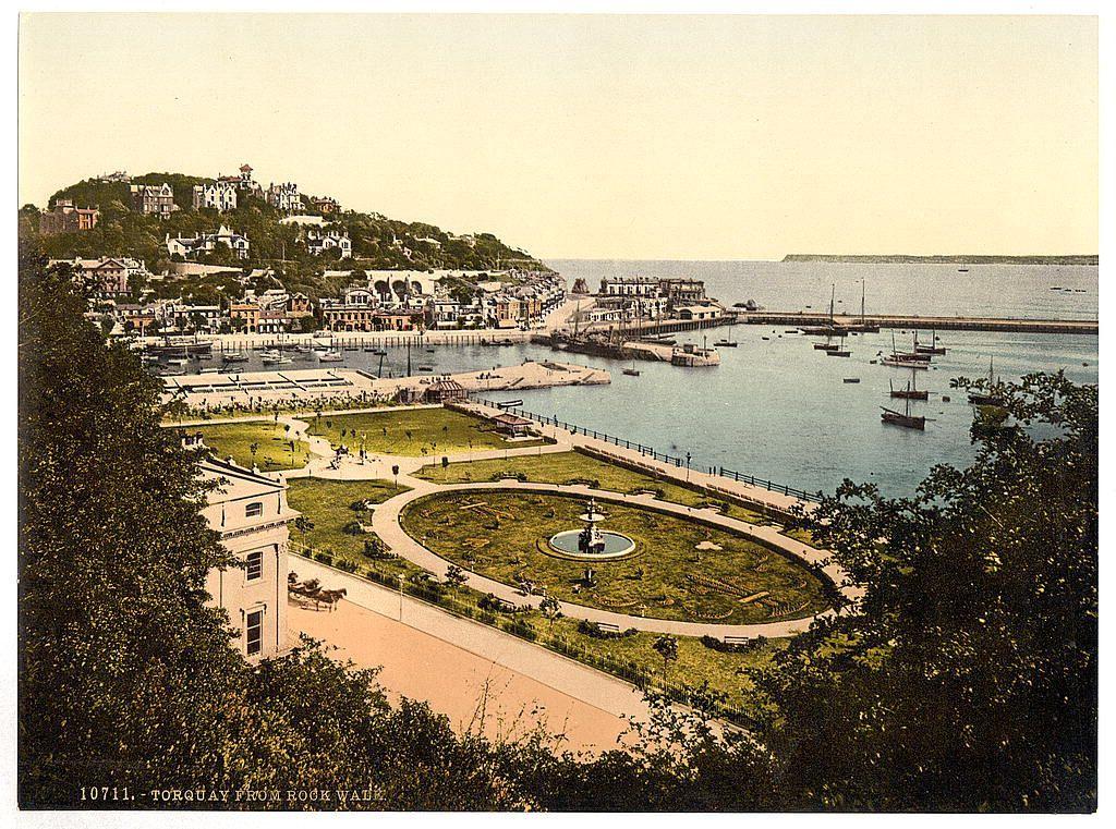 The image depicts a picturesque coastal town with various buildings and homes nestled on an elevated terrain overlooking the sea. In the foreground, there is a well-maintained garden featuring greenery, pathways, benches, and a central fountain, suggesting this might be a park or recreational area within Torquay. The coastline extends into calm waters where several boats are anchored at docks near what appears to be a harbor with larger vessels moored further out in the bay.

In the background, there is more architectural diversity ranging from small cottages to grander houses and possibly public buildings along the waterfront. A pier or breakwater juts out into the sea, enhancing navigation for ships entering the harbour. The coloration of this image suggests it's a vintage photograph with sepia tones typical of late 19th-century printing methods like photochroms.

The scene is serene and appears to be captured during daylight hours under clear skies, providing ample visibility across Torquay from Rock Walk, as indicated by the text at the bottom. The perspective seems high, possibly taken from a vantage point within or atop one of the buildings overlooking this scenic vista.