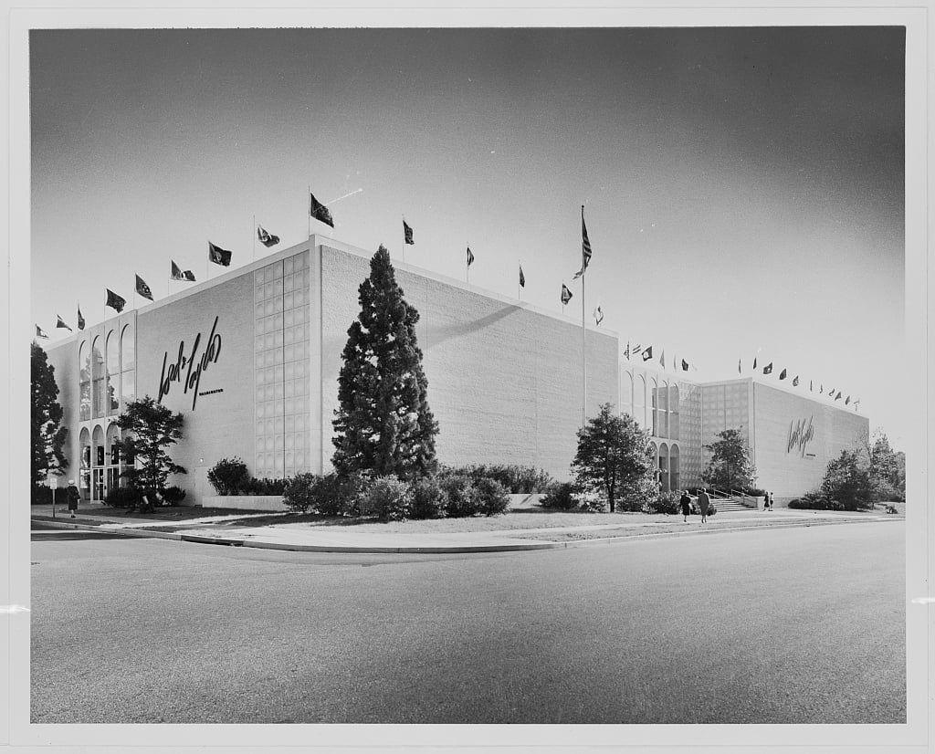 The image depicts a large, modern-style building with the name "Lord & Taylor" prominently displayed on its facade. The structure is characterized by clean lines and a simple architectural design, typical of mid-century commercial architecture. It features several rows of windows along the top portion and what appears to be multiple levels or floors within.

Various flags are mounted atop flagpoles positioned at different heights around the building's perimeter, indicating some form of international representation or affiliation for Lord & Taylor as mentioned in the context provided by "Lord & Taylor business in Washington, D.C. Western Ave., entrance from street 1959." The date associated with this image is specifically noted to be a year earlier.

In front of the building stands an evergreen tree and some landscaped bushes or shrubs provide greenery contrast against the stark white facade. A couple of individuals can be seen walking on what appears to be a paved path leading up to the entrance, suggesting that it was taken in daylight hours with enough natural light for clear visibility.

The photograph is presented as part of "Gottscho-Schleisner Collection," which seems to suggest an archival or historical significance. The source credits indicate ownership by the Library of Congress and provide a reference link for further information on this collection, along with s [...]