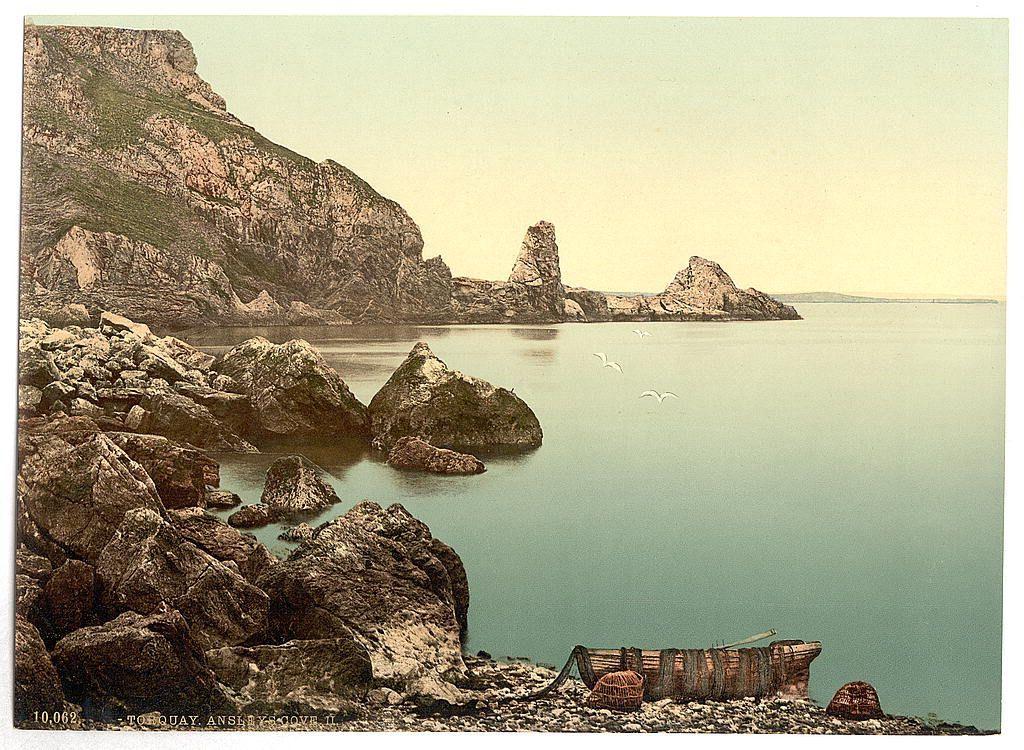 The image depicts a picturesque coastal scene with rocky outcrops and calm waters. A steep, rugged cliff rises dramatically from the sea on one side of the view, while several isolated rock formations break through the water's surface in the middle distance. The sky is overcast or has a pale yellow hue, suggesting an early morning or late afternoon time frame.

In the foreground, there are scattered rocks and boulders with small pools between them filled with clear blue-green waters reflecting some of the surrounding scenery. On these rocky shores rests what appears to be remnants of wooden structures, possibly old fishermen's boats or equipment storage areas, now in disrepair.

Two seagulls can be seen flying over the water, adding life and movement to the tranquil setting. The overall impression is one of natural beauty and perhaps a sense of isolation due to its remote appearance with no visible human activity aside from the remnants of what could have been local use or habitation.