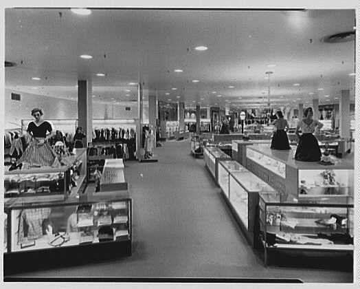 The image is a black and white photograph depicting the interior of what appears to be an old-fashioned department store. The view shows several wide aisles lined with merchandise, including clothing displays on racks along one side and various items showcased in glass display cases throughout the space.

On the left-hand side, there are two women standing behind a counter or serving area; they seem to be employees assisting customers. One is wearing an apron while attending to something on her workspace, possibly taking inventory or handling merchandise for sale. The other woman stands with hands clasped in front of her.

Further down the aisles, multiple sections contain racks displaying clothing items such as dresses and skirts. On either side, display cases exhibit a range of products that could include jewelry, accessories, and perhaps small home goods. 

The store has a spacious layout with tall ceilings supported by pillars or columns, suggesting an open floor plan typical for shopping centers during the mid-20th century. The lighting is bright, illuminating various items on sale.

In terms of context, this photograph was taken at Lord & Taylor's business in West Hartford, Connecticut, captured as part of a series showing different views within the store from May 25, 1953, by Gottscho-Schleisner Inc. The image is noted to be one negative on safety stock with dimens [...]