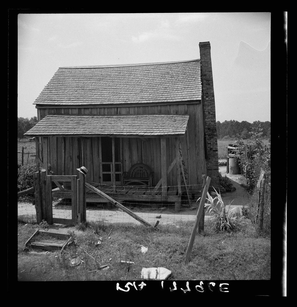 This black and white photograph depicts a rustic wooden structure with a steeply pitched roof, indicative of traditional building methods possibly from the early to mid-20th century. The house is characterized by its weathered appearance, suggesting age or neglect. It has a prominent gabled front section and appears to be constructed primarily using vertical plank siding. A small chimney on the right side hints at an interior fireplace for heating.

The dwelling features double-hung windows with visible sills and a central door that seems sturdy but worn out over time. The entrance is partially obscured by what looks like a wooden bench or step, indicating limited space around it. To the left of this structure stands another building attached via a small enclosed passageway; its purpose isn't immediately clear.

In front of these buildings, there's an assortment of debris and discarded items scattered on the ground near the fence line, including what appears to be gardening tools or yard waste, suggesting sporadic maintenance. A tall straw hat lies in the foreground, possibly belonging to a worker or resident not currently visible within the frame. The overall setting is rural with overgrown grasses hinting at a lack of recent human activity.

The photograph carries an air of desolation and abandonment, emphasizing solitude rather than domestic life. It captures a moment  [...]