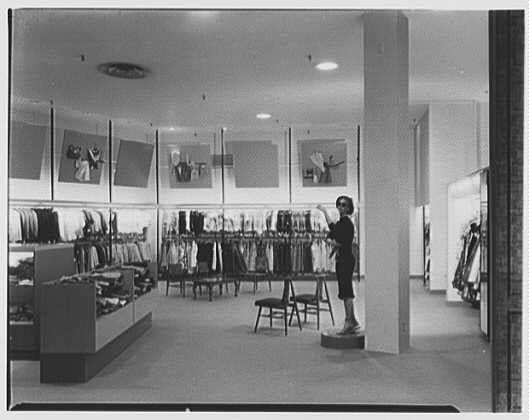 The image depicts an interior view of a clothing store from the mid-20th century. A female salesperson is standing on a stool, arranging merchandise near one wall filled with various garments hung up for display. The room has bright lighting and features rows of shelves stocked with clothes as well as racks lined against the walls. On the ceiling are mounted spotlights that illuminate specific areas within the store. In the background, several large framed posters or advertisements hang on the back wall showcasing women in different outfits. A few chairs can be seen around a small display table near the foreground. The overall atmosphere suggests an organized and well-lit retail space with emphasis placed on presenting clothing items to potential customers.