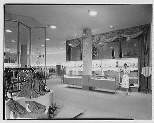 The image is a black and white photograph of an interior scene, likely from the early 1950s given its vintage appearance. It depicts what appears to be a department store or boutique named "Fantasia shop," as indicated by text at the bottom of the photo.

Within this setting, there are various displays featuring clothing items on mannequins and racks along with jewelry showcased in glass cases against the wall. The overall layout is spacious, well-lit, and organized for customer viewing. A female store employee or shopper can be seen walking through the aisle towards the right side of the frame.

The lighting fixtures overhead suggest a contemporary design typical for that era. Curtains adorned with floral designs add an elegant touch to the decor along one wall, further enhancing the sophisticated atmosphere of this shopping establishment.