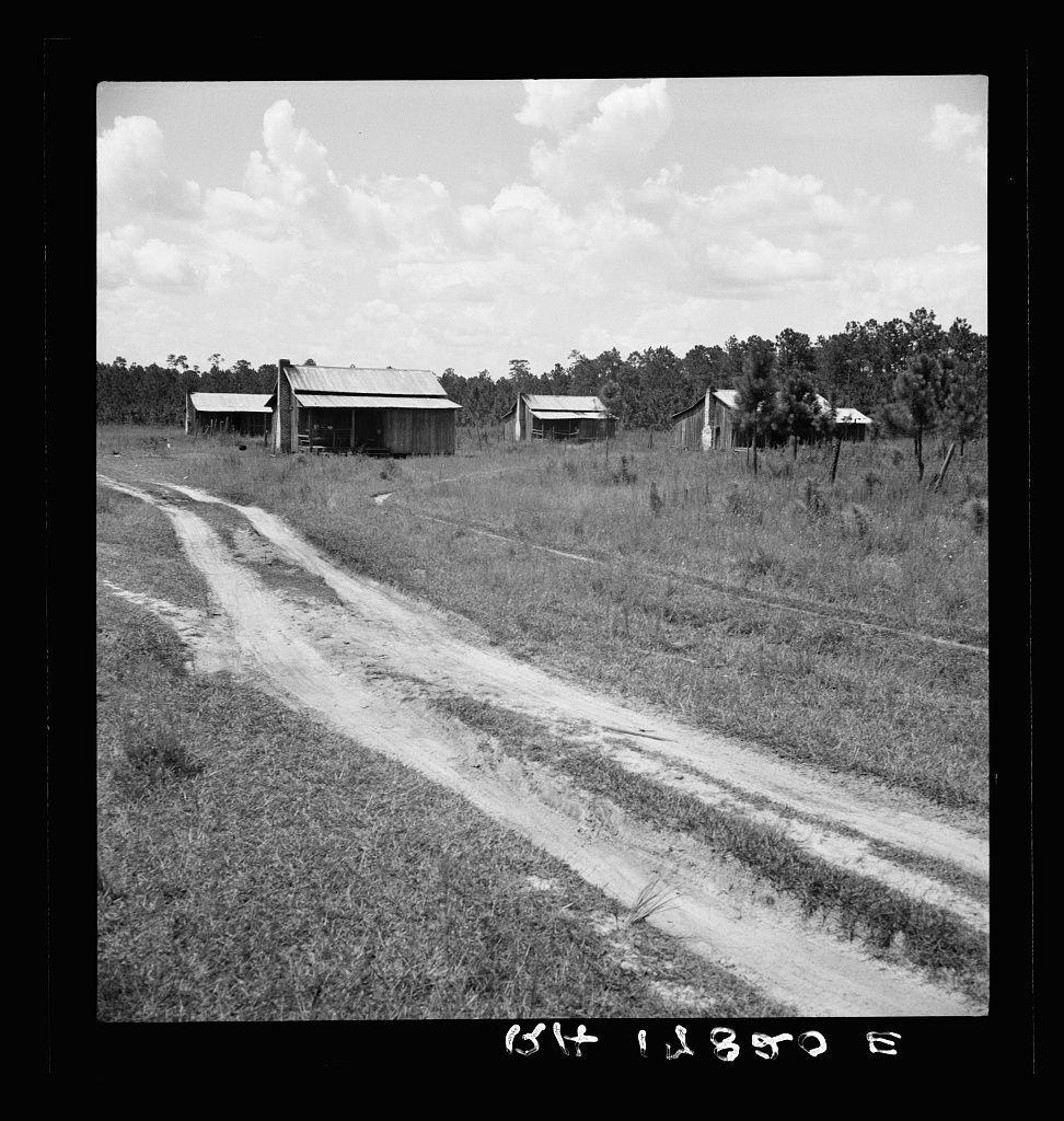 The image is a black and white photograph depicting an expansive rural landscape with sparse buildings. A dirt road, which seems to be unpaved, cuts through the middle of the frame from bottom left to top right, creating a diagonal line that draws attention into the distance.

On either side of this central pathway lie what appear to be dilapidated structures; these may be old sheds or small barns typical of agricultural settings. The buildings are simple with basic roofing and lack modern amenities like siding. Their state suggests they have been exposed to weathering for a considerable time, perhaps indicating an abandonment or disuse.

The surrounding area is open field dotted sporadically with tall grasses or weeds, hinting at neglect in terms of cultivation but also suggesting that this land could be used for grazing livestock due to its overgrown nature. The vegetation appears wild and untamed rather than managed crop fields.

In the background stands a dense collection of trees, likely part of a forested area on the periphery of what's otherwise an open field or scrubland. Their presence adds depth to the scene but also contributes to an overall impression that this setting is remote and possibly rural.

A date stamp in white text at the bottom right indicates "1936," which places it within the context of early 20th-century American history, a period known for its  [...]