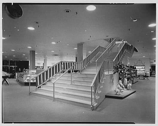 The image depicts an interior view of a mid-20th century store, likely from the early 1950s. The focus is on a large staircase with metal railings and steps that lead up to another level within the building. Below the stairs, there are displays showcasing mannequins dressed in fashion attire, suggesting this could be a department or retail store.

The architecture features numerous recessed ceiling lights creating a well-lit environment. The flooring is not entirely visible but appears to be of a light color, possibly terrazzo or polished concrete. A few other elements can be seen around the space: tables that might hold products for sale and additional shelving units stocked with merchandise in various areas.

The absence of windows indicates an enclosed structure, perhaps located within an urban shopping center or mall setting. The image is monochromatic, which suggests it's a black-and-white photograph from its time period.

This specific photo may be associated with Lord & Taylor, as mentioned in the context provided. This department store chain was active during that era and known for various locations across cities like Hartford, Connecticut (Westfarms Mall). The date referenced is May 25th of an unspecified year within this timeline (likely around or before WWII due to its design elements).

The source credits Gottschol-Schleisner Inc., a company noted as "1 negati [...]
