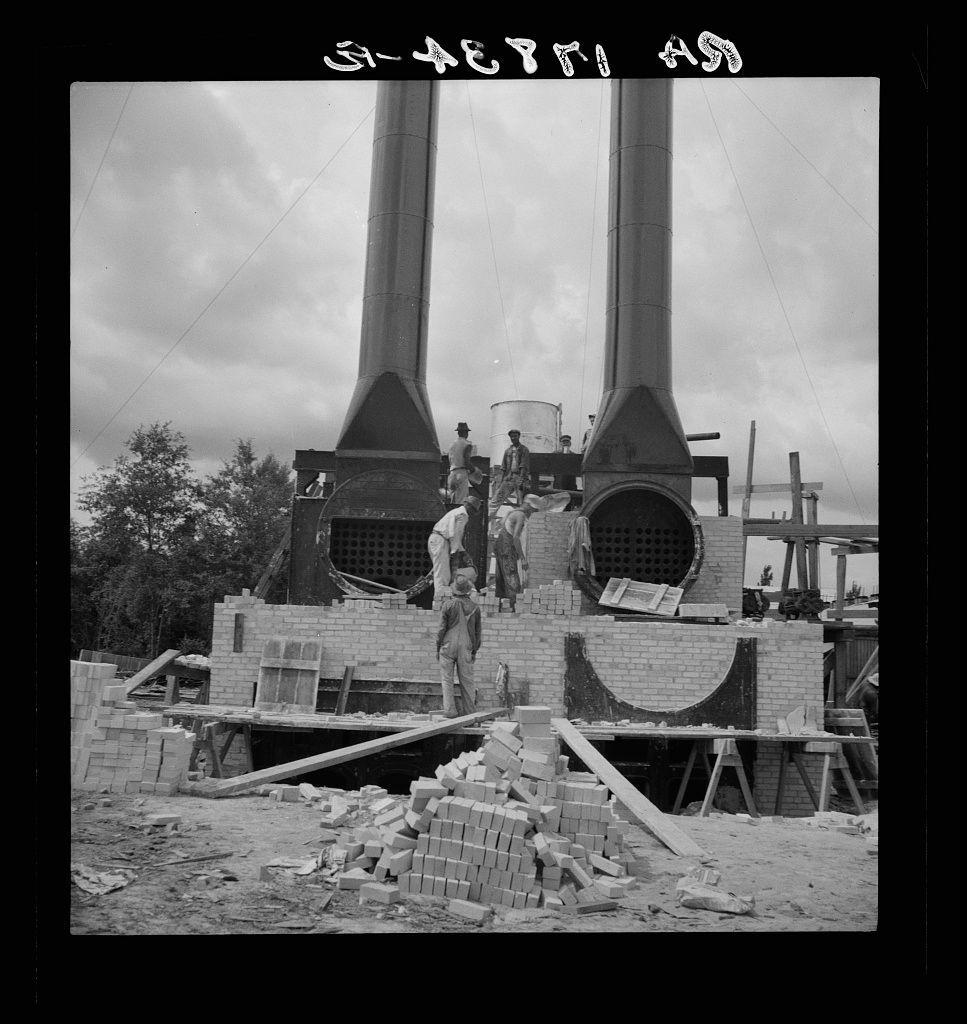 The black and white photograph depicts a construction site where several individuals are working on what appears to be the assembly or maintenance of large industrial structures, specifically chimneys. The most prominent features in the image are two tall concrete smoke stacks with cylindrical bodies topped by conical flues.

In front of these towers is an incomplete brick structure, possibly part of their foundation or support system. A pile of bricks lies haphazardly on the ground to one side, and several wooden supports can be seen scattered around the site for stability during construction work. 

The workers are engaged in various activities; some stand atop the completed sections of the chimneys while others appear to be handling materials or assessing their progress.

In the background, trees suggest that this industrial facility is located near a forested area. The sky is partly cloudy, indicating fair weather conditions at the time of photography.

There's handwritten text on top in Hebrew: "Keren Kayemah," which translates from Hebrew as 'Great Harvest,' likely referencing the name or purpose related to the construction activity captured within this photograph.