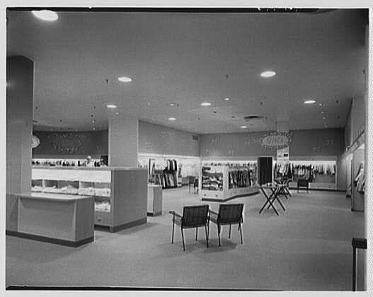 This black and white photograph shows the interior of a department store, likely from around 1953. The floor is tiled with large square tiles, and there are several display cases along one wall filled with what appears to be coats or outerwear. Another row displays more items on racks against another wall.
In the center area, there's an open space with three black chairs arranged in a triangle formation facing towards the middle of the room where other merchandise is displayed. The ceiling has recessed lighting fixtures installed at regular intervals throughout the store. There are no visible signs or labels that provide additional context about what kind of items are being sold.
The overall atmosphere appears to be clean and modern for its time, with an emphasis on presenting products in a clear view without much clutter.