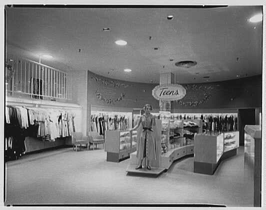 The image shows an interior view of a clothing store, specifically the Teen's section. The photo is in black and white, suggesting it was taken during a time when color photography wasn't widely used or accessible.
In the center stands a mannequin dressed elegantly in a long dress with gloves on its hands. Above the mannequin hangs a large sign that reads "Teen's" accompanied by additional text advertising specific clothing items such as blouses, sweaters, coats, and pants. The store has an open layout with racks of clothes displayed along the walls; some garments are hung neatly while others appear folded.
There is shelving in front of the mannequin housing various accessories or small apparel pieces. To the left side of the photo, there's a rail extending across part of the room filled with more clothing items for sale. Overhead lighting illuminates the store, and there seems to be an escalator or stairs leading up from this area.
The setting appears tidy and well-organized, aiming at creating an appealing shopping environment for young customers browsing teen apparel options during the mid-1950s in Connecticut as indicated by Lord & Taylor business reference.