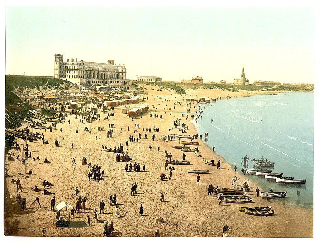 This image depicts a bustling beach scene from Long Sands in Tynemouth, England. The photograph captures the essence of late 19th-century seaside leisure with numerous people scattered across the sandy shore engaged in various activities such as walking, sitting on benches, and playing games or cards. Boats are anchored along the shoreline, some closer to the water's edge while others further up the beach. There is a significant concentration of visitors near what appears to be a large pavilion-like structure with multiple stories and rows of windows, likely serving as a seaside resort or public building.

The background features rolling sand dunes leading towards more densely populated areas of Tynemouth with several buildings that hint at residential or commercial purposes. The skyline is punctuated by a notable church spire to the right, indicating a mix of secular and religious architecture within this coastal town. The photograph has a vintage sepia tone indicative of its age, possibly dating between ca. 1890 and ca. 1900.

The overall impression conveyed through the image is one of leisurely recreation in an established seaside resort setting during what was likely a popular period for such activities on British shores.