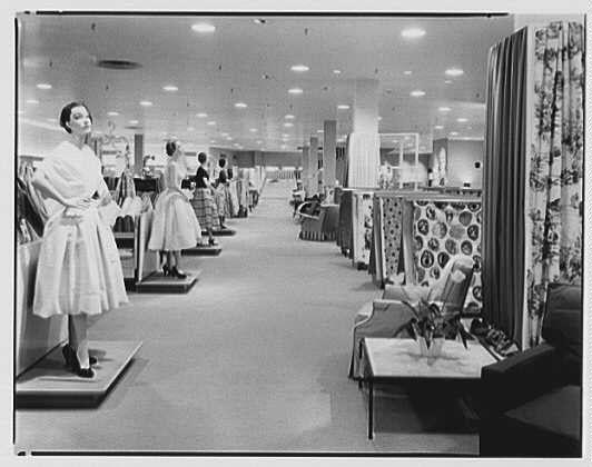 The image depicts an interior view of a retail store, specifically focusing on the fashion and home decor sections. A long row of mannequins is lined up along one side, each dressed in different styles of mid-century modern women's clothing including skirts, dresses, coats, and accessories such as hats and gloves.

On display are various patterns and textures for upholstery fabrics; these include floral designs with large circles and other geometric shapes. The floor appears to be a polished concrete surface typical of the era, while seating arrangements feature patterned sofas and chairs that complement the overall decor theme.

The environment is well-lit by overhead fluorescent lights which reflect off the shiny surfaces within the store. Curtains are visible in the background near one side, possibly delineating different sections or aisles for customers to browse through.

This scene captures a moment from 1953 at Lord & Taylor business located in West Hartford, Connecticut; this specific image is attributed to Gottschalk-Schleisner Inc., indicating it may be part of their collection. The overall atmosphere evokes the mid-century modern aesthetic with its clean lines and love for patterns and textures.