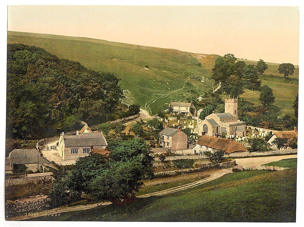 The image depicts a picturesque rural landscape of the village of Upwey in Dorset, England. It is an elevated view showing winding pathways and lush greenery enveloping quaint stone buildings with thatched roofs typical of English countryside architecture from around 1890 to 1900.

In the foreground, there's a serene pond reflecting its surroundings, flanked by neat hedges leading up to cobblestone paths. The middle ground is dominated by traditional village structures including houses and what appears to be an old church with a prominent tower featuring clock faces on each side visible against clear skies during daylight hours.

The background reveals gently rolling hills covered in rich green vegetation stretching into the distance, suggesting that this could possibly represent early 20th-century England. The image is marked as being part of the Flickr Commons project by Loener NL and has an identifier number indicating its cataloging within a larger collection of historical images from around the world.

The overall atmosphere conveyed through the colors ranging from vibrant greens to earthy tones evokes tranquility, simplicity, and nostalgia for a bygone era.