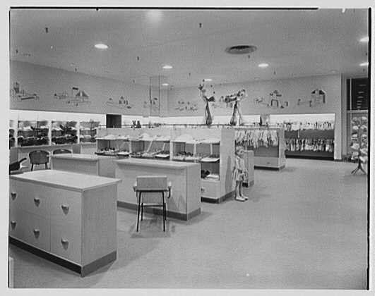 The image shows an interior view of a retail store, likely from the mid-20th century based on its style. The black and white photograph captures a spacious department with multiple counters or checkout lines where customers can make purchases. A woman is standing near one counter, possibly engaging in shopping activities. Above each counter are shelves displaying various items for sale, which appear to be clothing given their appearance.

The store has an open layout with several light fixtures on the ceiling providing illumination. The floor seems clean and well-maintained, contributing to a neat environment suitable for commerce. In one corner of the room, there is what appears to be a tall display case or stand showcasing additional merchandise. A chair is placed near this area.

The walls are adorned with decorative elements that seem like stylized depictions of boats or ships, possibly serving as wall art or part of an ornamental design theme for the store's interior decor. The overall atmosphere suggests a period where retail shopping was becoming more organized and consumer-friendly compared to earlier times in history when such establishments were simpler affairs.

The photograph is credited to Gottschol-Schleisner Inc., suggesting that this image may be from their collection, which specializes in documenting various aspects of the early-to-mid 20th-century period [...]