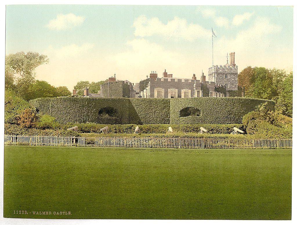 The image depicts a historic castle named Walmer Castle, located in Kent, England. The photograph appears to be from around the late 19th or early 20th century, based on its style and coloration which suggests it's an old postcard. It shows the fortress-like structure with multiple towers topped by conical roofs and a flag flying atop one of them. A series of archways are visible through what looks like a high hedge that encloses part of the castle grounds.

In front of this, there is a neatly manicured lawn extending towards a wooden picket fence which separates it from an area with more greenery and trees in the background. The sky above is partly cloudy but bright, indicating good weather at the time the photo was taken. There are also sheep grazing on what appears to be grassy ground behind this hedge.

The overall scene gives off a serene historical ambiance, typical of English country estates during that era.