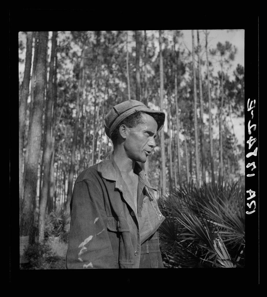 The image is a black and white photograph depicting an individual standing outdoors. The person appears to be wearing casual work attire, including a hat that seems suited for sun protection and possibly indicative of laborious outdoor activities like farming or forestry maintenance. Their jacket has visible stains, suggesting active use in dirty tasks.

Behind the subject, there's a background consisting mostly of tall trees with thin trunks, which could suggest a pine forest setting, often associated with turpentine production as mentioned in the provided context reference to "Turpentine 'chipper' near Homerville, Georgia." The vegetation includes some darker, bushier plants that may be indicative of underbrush commonly found around such areas.

The photograph has text overlaid on its borders which is likely date-related or descriptive information, common with historical photographic prints. However, the exact meaning and context are not clear without further interpretation beyond what's provided in this brief description. The overall mood conveyed by the image seems to be one of contemplation or respite within a working environment.