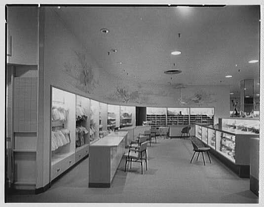 The image depicts an empty department store interior from a past era, specifically the ladies' clothing section labeled "Layette" dated May 25, 1953. The black and white photograph captures several display cases filled with garments on either side of a central aisle that extends toward the back wall. In between these displays are rows of empty chairs for shoppers to sit. The ceiling is adorned with recessed lighting fixtures in an organized pattern.
The store appears clean, well-organized, and inviting, typical of mid-century retail design aesthetics. There's no visible branding or signage indicating a specific location beyond "Layette," which could be the name of this particular section within Lord & Taylor stores across various locations during that time period. The photograph is credited to Gottschalk-Schleisner Inc., suggesting they were responsible for taking and possibly producing it as part of their business activities.
The image serves as a historical snapshot, showcasing the store's interior design elements such as flooring materials, display cases' shapes and sizes, furniture styles including chairs and counters, lighting fixtures on both the ceiling and walls. It captures an atmosphere that would have been familiar to shoppers during this period in Connecticut or other locations where Lord & Taylor operated.
This photograph can be found under reference number " [...]