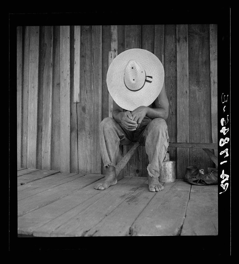 The black and white photograph depicts a seated individual wearing casual attire consisting of long pants, an open-collar shirt, and no shoes. The person is sitting with their legs crossed at the ankles on what appears to be wooden planks or flooring. They are donning a large straw hat that obscures most features except for eyes and nose peeking through the brim's edges. One can observe various objects in close proximity: boots placed beside them, shoes positioned across from one another, a small bucket near their right foot, which is resting on it, and what appears to be a tin or container with metal elements on the left side of this person’s feet.

The background consists of vertically aligned wooden planks that form part of a wall. On the upper-right corner, there's text overlaid in white characters against a black border: "21-8731 AS" and beneath it, additional text possibly related to identification or cataloguing, with some indecipherable symbols.

The setting seems rustic, indicative perhaps of rural living conditions commonly associated with manual labor jobs like turpentine production. The image has an air of contemplation or waiting as the person sits in solitude on a porch-like structure.