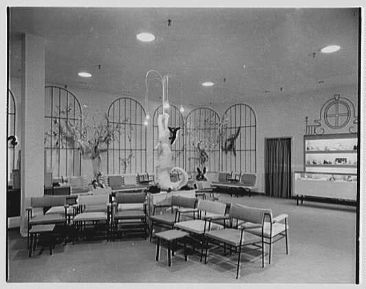 The image depicts an interior scene, likely from a vintage department store or boutique. It shows rows of seated furniture including chairs and sofas arranged in neat lines on what appears to be carpeted flooring. Large arched windows dominate the back wall with decorative items displayed within them.

Centerpiece of this setting is an ornate sculpture resembling a flower, possibly made of metal, standing prominently amidst several statues or sculptures that seem like figures from mythology or classical themes. These are dispersed throughout the room on stands and in niches.

To one side, there's a display case with more items for sale, suggesting the space might be part of a jewelry store within this larger venue. The lighting is soft but ample, casting gentle shadows around the sculptures and seating arrangements.

Overall, it has an elegant and inviting ambiance typical of mid-century retail design where presentation was key to attracting shoppers' attention.